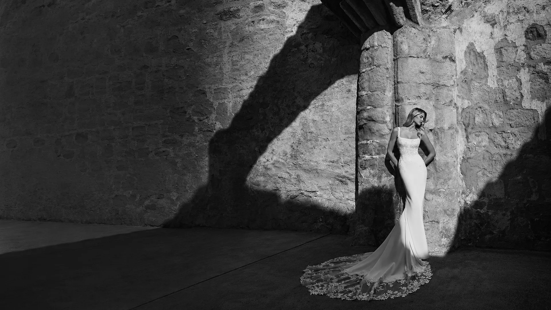 Bride in a fitted lace and crepe wedding dress with a long train, photographed in black and white against a sunlit stone wall — BOE Bridal, Eccleshall