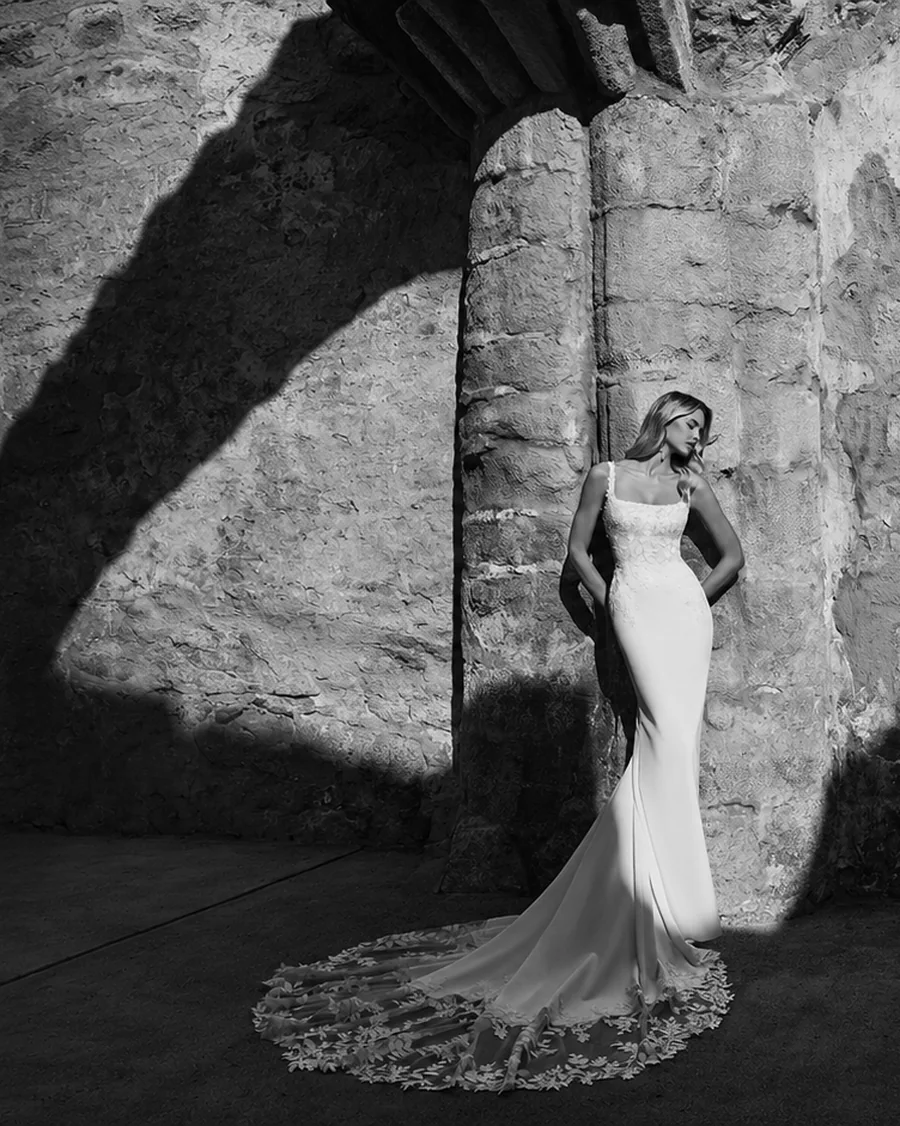 Bride in a fitted lace and crepe wedding dress with a long train, photographed in black and white against a sunlit stone wall — BOE Bridal, Eccleshall